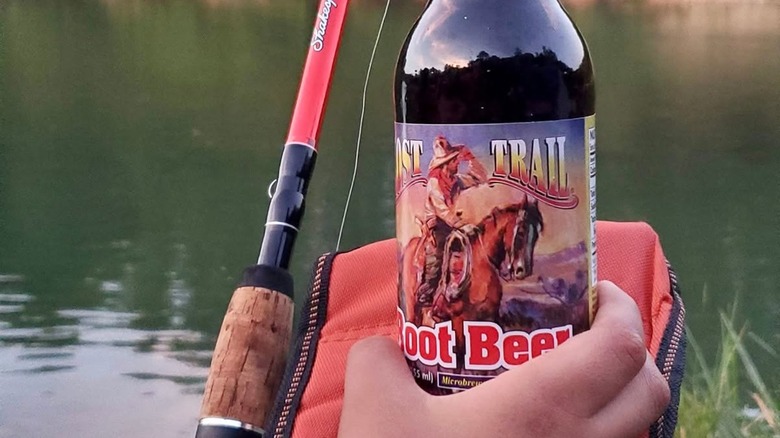 Hand holding a bottle of Lost Trail root beer with a fishing pole and river in the background