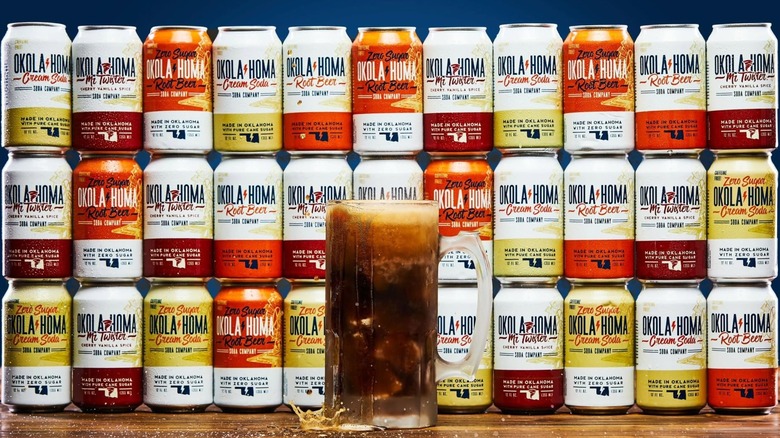 Mug of root beer with a wall of assorted Okola-Homa soda cans in the background