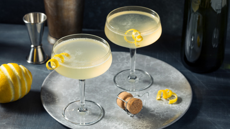 Two coupe glasses filled with French 75 and garnished with lemon twists on a platter beside cocktail-making paraphernalia