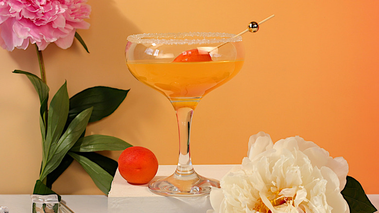 Orange-colored cocktail in a coupe glass garnished with an apricot on a cocktail pick