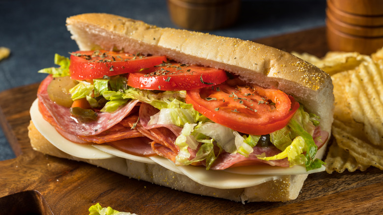 An Italian Sub sitting on a wooden board with a pile of ruffled potato chips behind it