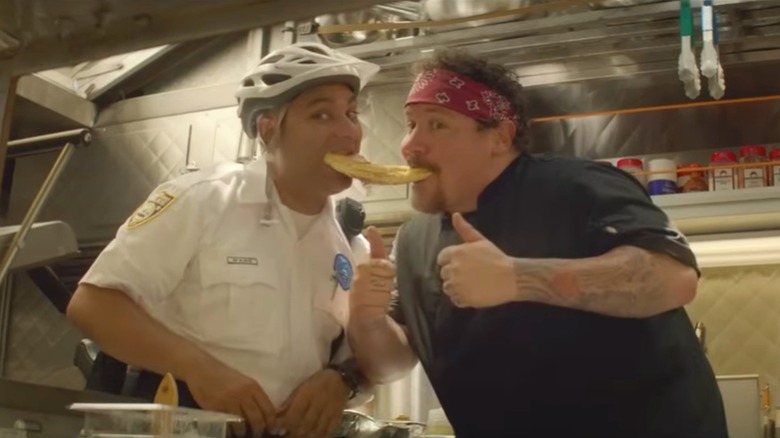 A still from "Chef" in which Carl and a man in a bicycle helment hold a piece of bread in their mouths inside the food truck