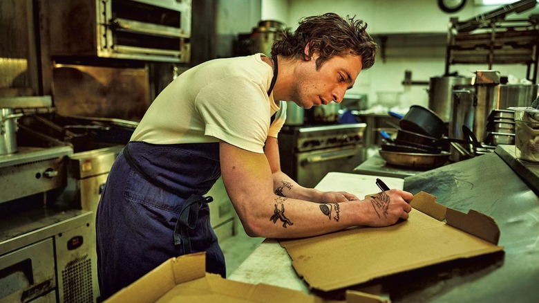 Carmy from "The Bear" wearing an apron and taking notes on a pizza box in the kitchen of The Beef