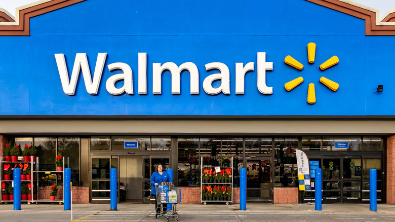 The exterior of a branch of Walmart, with an employee pushing a shopping cart from its doors