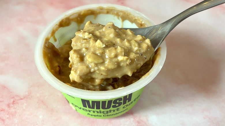 Spoonful of MUSH apple cinnamon oats over container of oats on pink marble surface