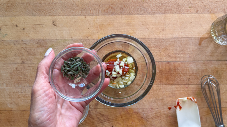 Hand holding small bowl of chopped rosemary over bowl with garlic and tomato paste
