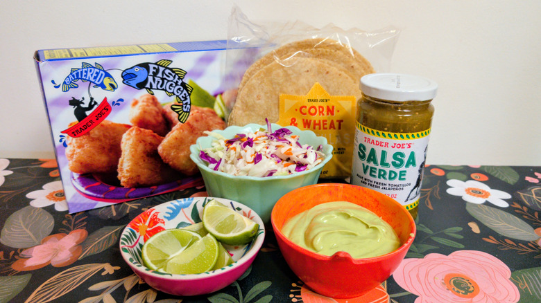 Trader Joe's Battered Fish Nuggets, Corn & Wheat Tortillas, and Salsa Verde on a counter next to bowls of lime wedges, cabbage slaw, and avocado crema