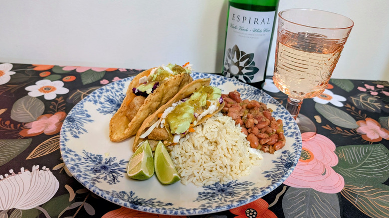 A plate with two fish tacos, lime wedges, rice, and beans next to a bottle of Espiral Vinho Verde and a glass of wine