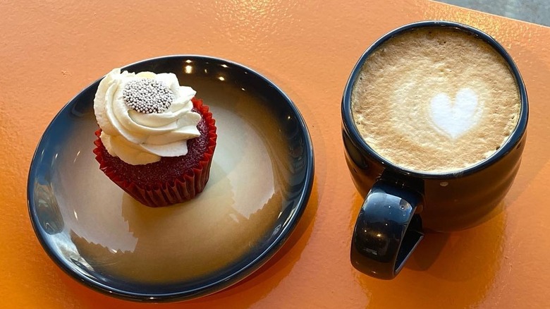 Red velvet cupcake topped with swirled whipped cream on a plate next to coffee