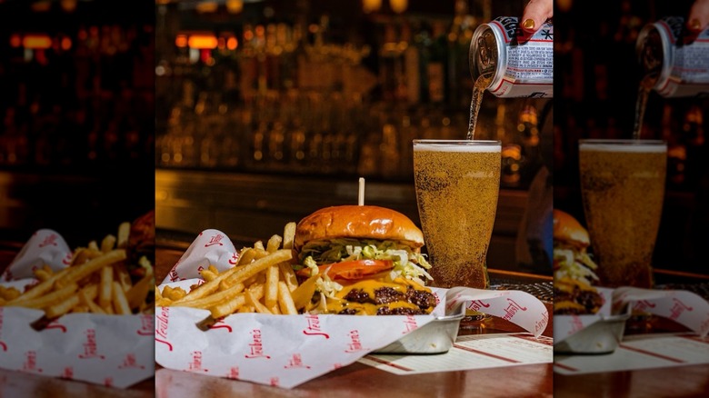 Can of PBR being poured into a fizzing pint glass next to a burger and fries sitting on parchment paper in a basket