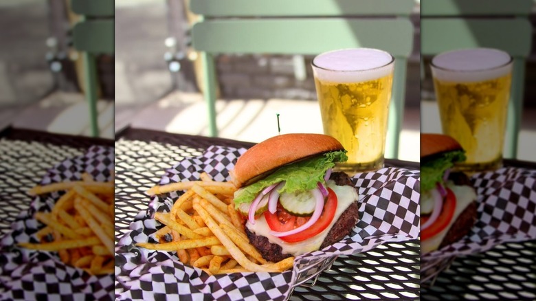 Burger sitting on checkered paper in a basket with fries next to a draft beer on an outdoor table