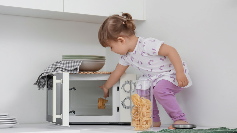 Toddler standing on a counter, putting dry pasta into a microwave oven