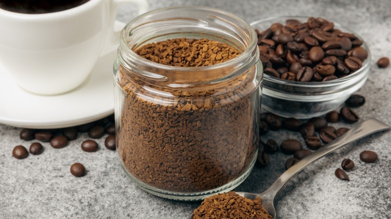 A glass jar of instant coffee with a spoon, coffee beans, and a mug of coffee