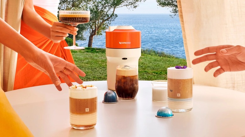 Hands reaching for various coffee drinks next to an orange Nespresso machine near the ocean