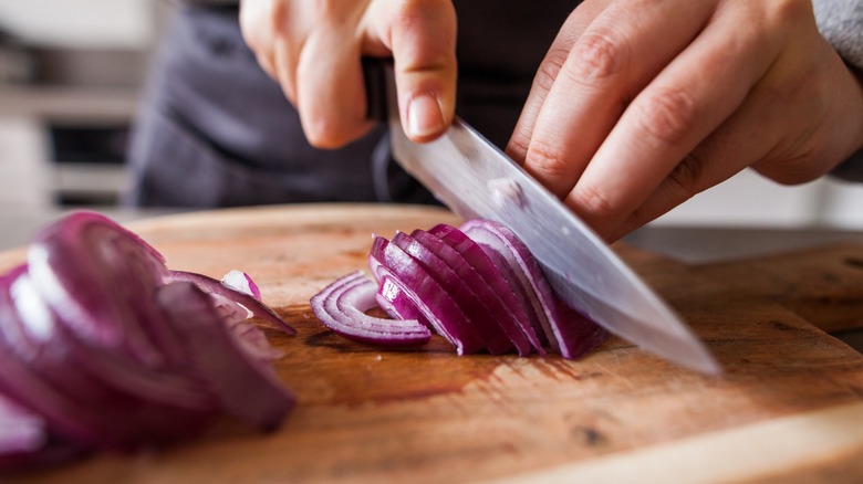 someone's hands using a chef's knife to slice red onions on a cutting board
