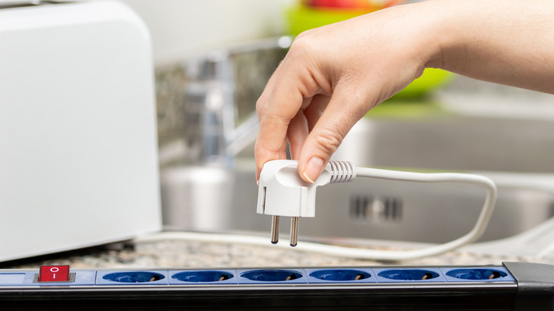 A close-up of a woman's hand plugging an appliance cord into a power strip in a kitchen