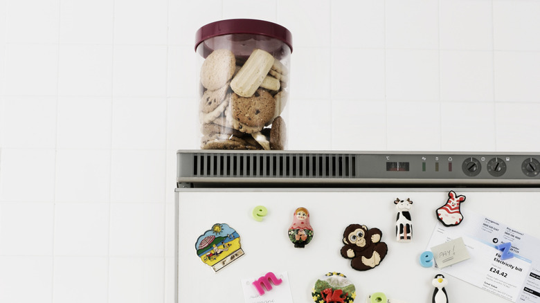A clear jar of cookies stored on top of a fridge covered in colorful magnets.