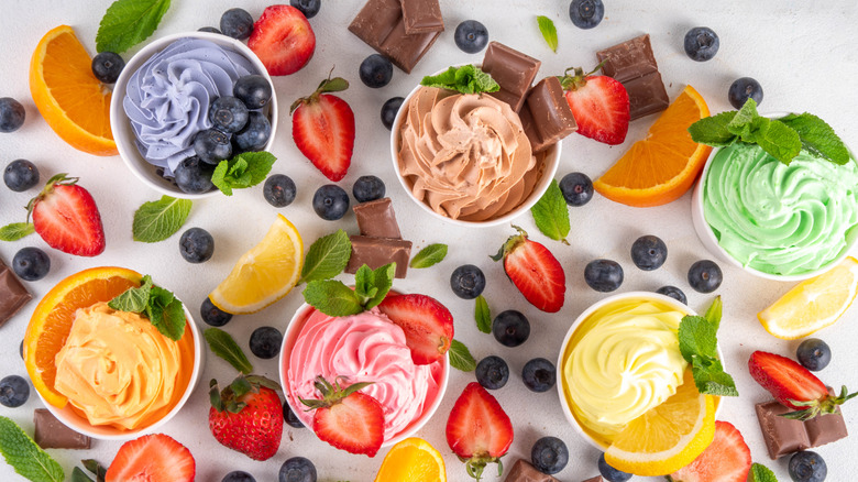An assortment of flavored yogurt in containers with blueberries, oranges, strawberries, chocolate pieces, lemon slices, and mint leaves around them on the table.