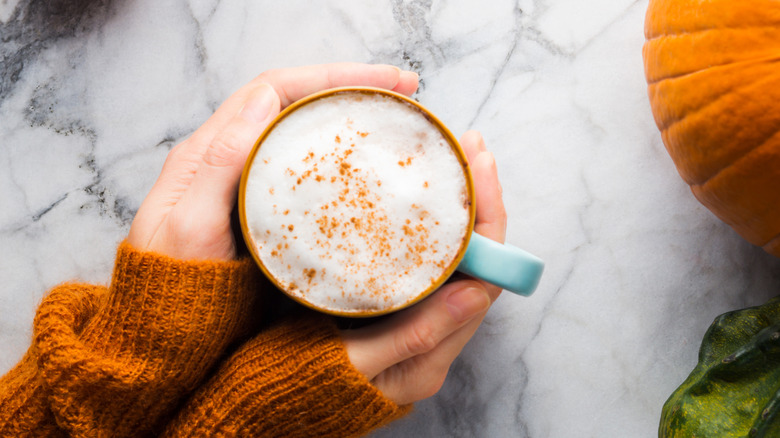 Hands covered by sweater holding a cup of coffee with pumpkin in frame