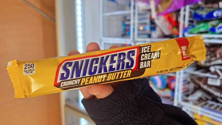 Hand holding a Snickers Crunchy Peanut Butter Ice Cream Bar in a store's frozen snacks section