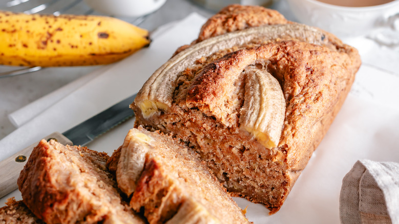 A loaf of banana bread with bananas cooked into the top, sliced on a table