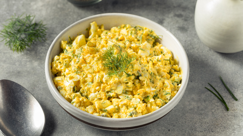 A bowl of egg salad is set on a counter with dill and a spoon.