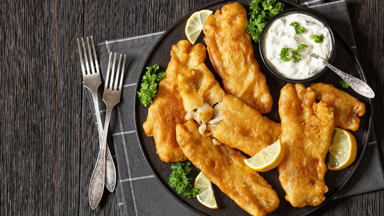 black plate of fried fish loins with tarter sauce cup, parsley, napkin, forks