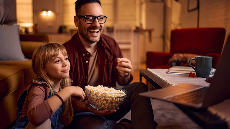 Father and daughter eating popcorn and sitting on a living room floor laughing and watching a laptop