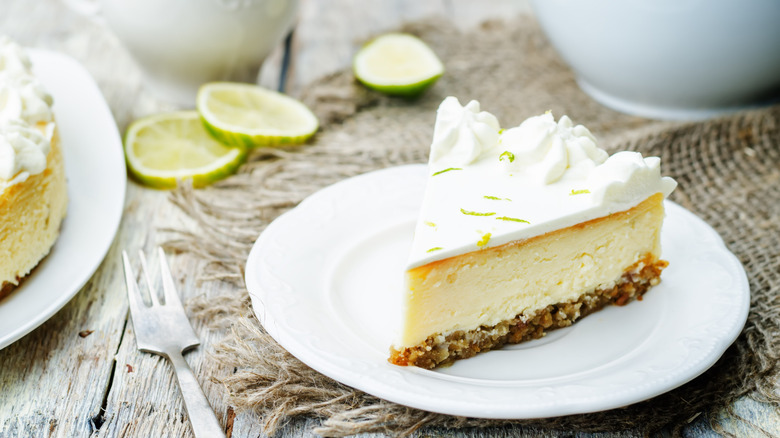 A slice of key lime cheesecake on a plate with slices of limes in the background.