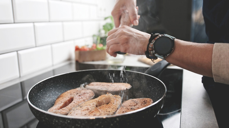 A person salts salmon steaks frying in a pan