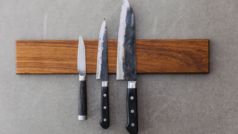 Three different sized knives hanging on a magnetic strip on the wall of a kitchen