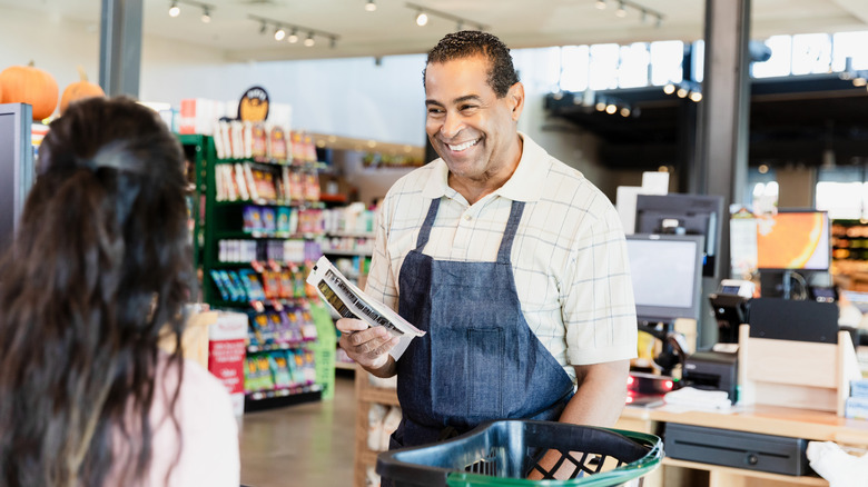 A smiling male cashier wearing a denim-colored apron speaking to a female customer
