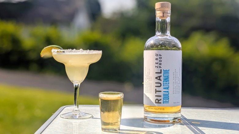 Bottle of Ritual tequila with a full shot glass and a margarita on a white table outside