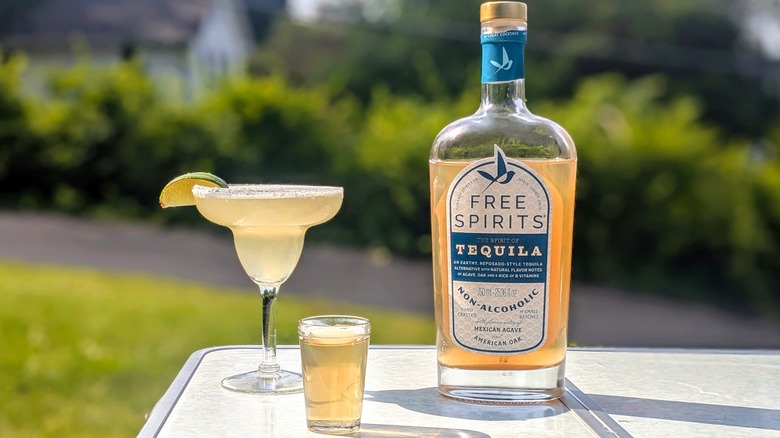Bottle of Free Spirits tequila with a full shot glass and a margarita on a white table outside