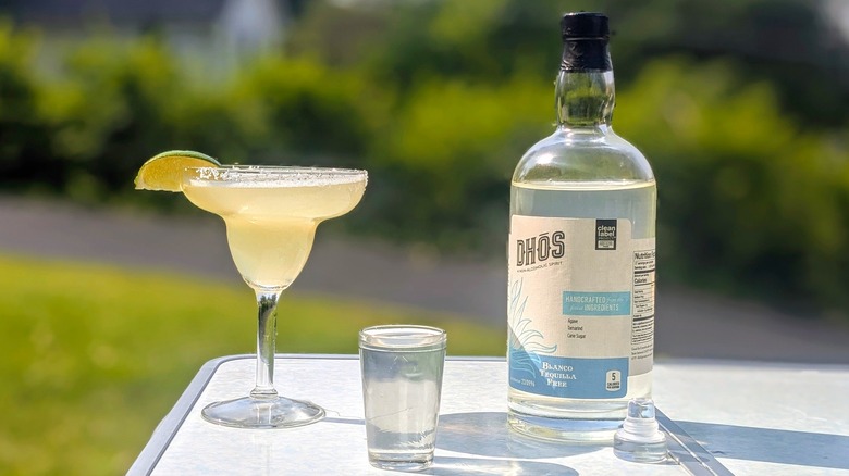 Bottle of Dhōs tequila with a full shot glass and a margarita on a white table outside