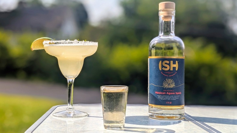 Bottle of ISH tequila with a full shot glass and a margarita on a white table outside