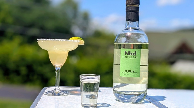 Bottle of Nkd tequila with a full shot glass and a margarita on a white table outside