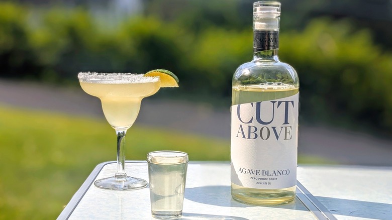 Bottle of Cut Above tequila with a full shot glass and a margarita on a white table outside
