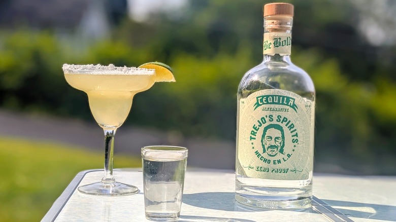 Bottle of Trejo's Spirits tequila with a full shot glass and a margarita on a white table outside