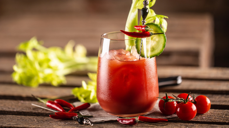 Bloody Mary cocktail in glass garnished with cucumber, hot peppers, and celery next to peppers and cherry tomatoes