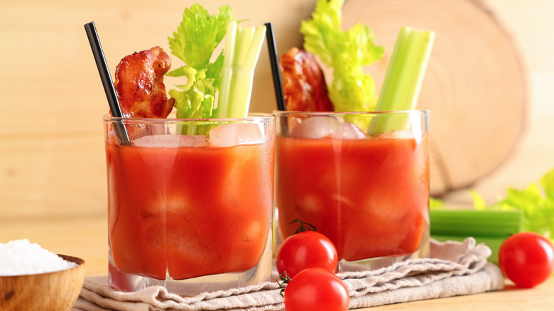 Two short glasses of Bloody Mary cocktail with black straws, bacon strips, and celery garnishes