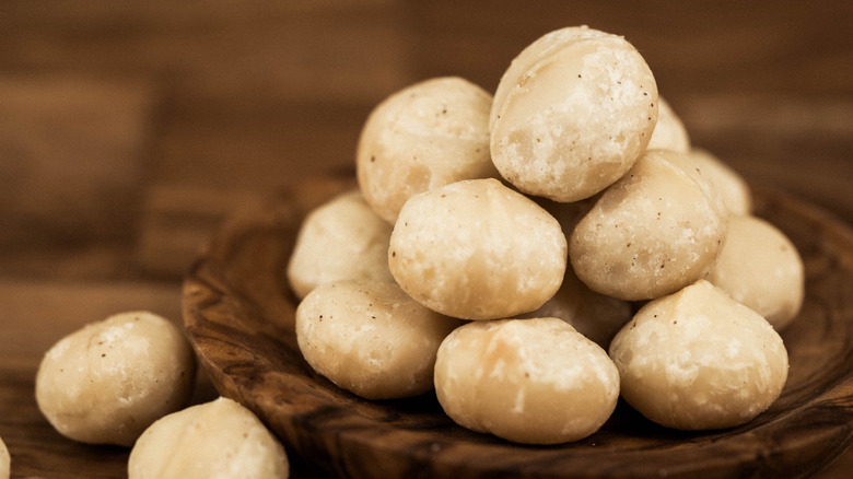 a small wooden bowl of macadamia nuts surrounded by additional nuts on a wooden table