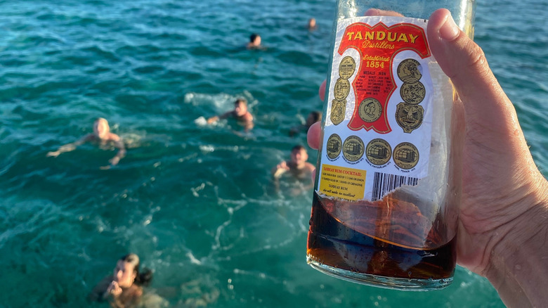 person holds Tanduay rum bottle above ocean with swimmers