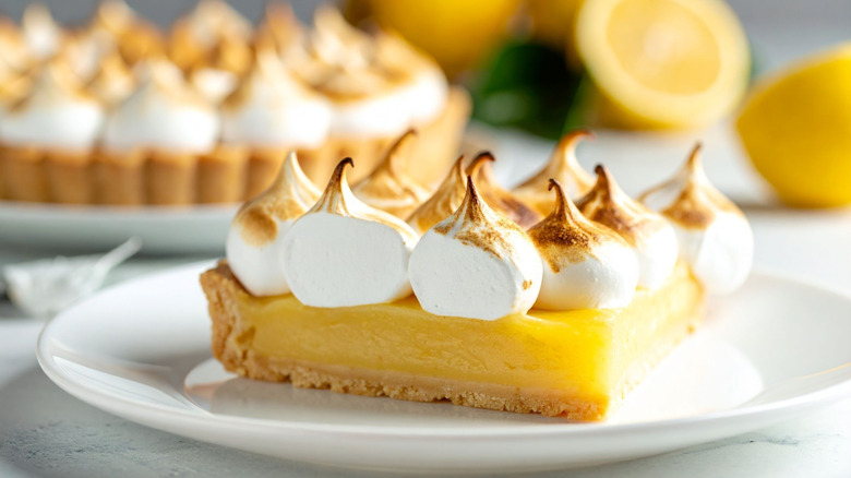 A slice of lemon meringue pie on a plate with the rest of the pie and lemons in the background