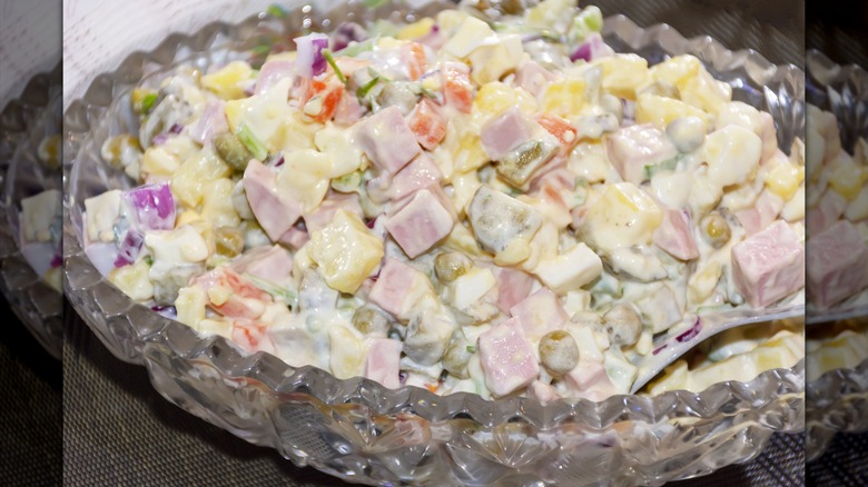 A glass bowl of salad Olivier with ham, vegetables, and potatoes.