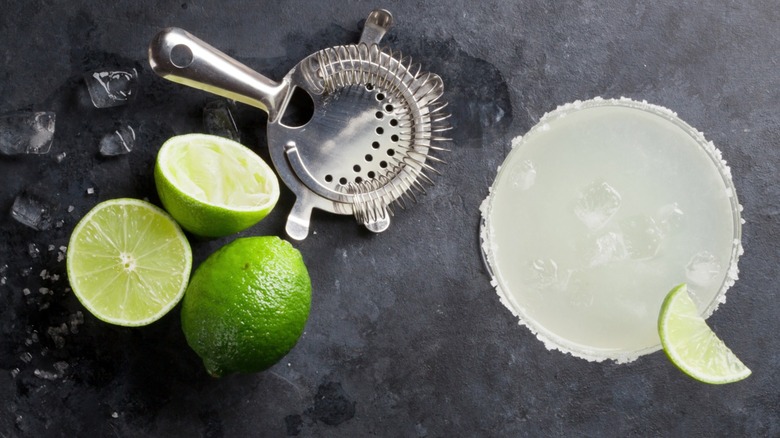 Aerial view of a margarita on the rocks with limes, ice, and tools