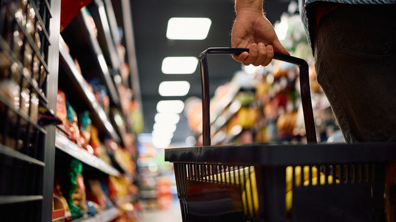 A close up of a shopper holding a basket while walking down an aisle at a supermarket