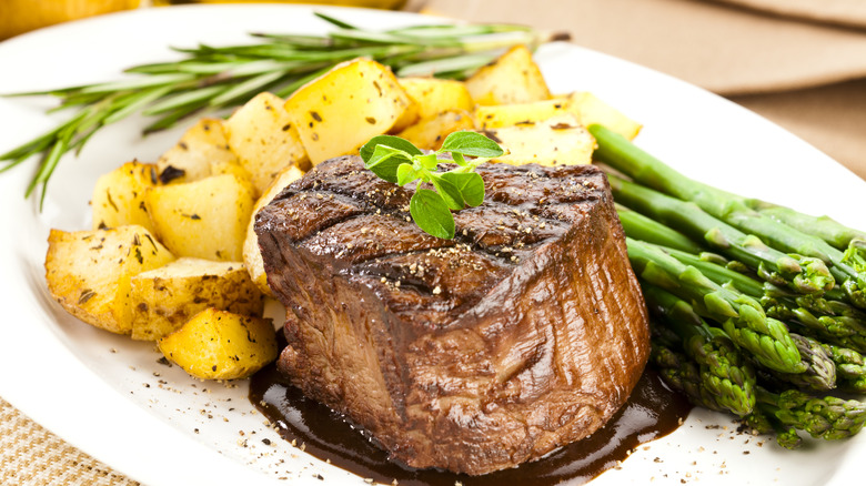 A filet mignon on a white plate with roasted potatoes and asparagus, garnished with a sprig of rosemary