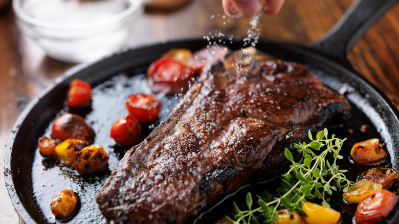 A New York strip steak cooking in a cast iron skillet, garnished with sea salt, cherry tomatoes, and watercress