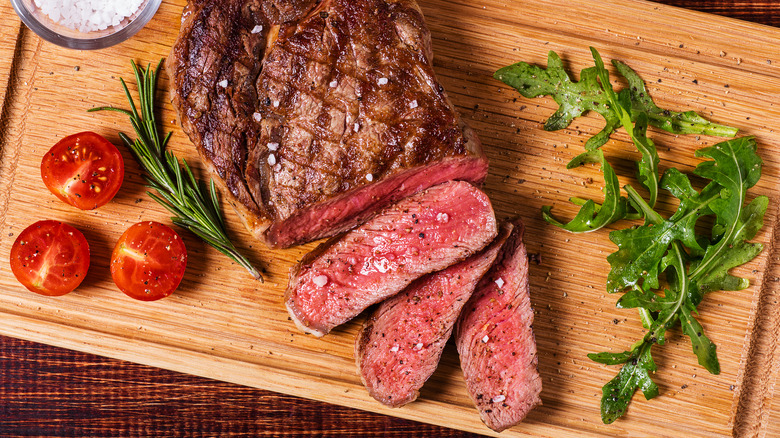 A large piece of ribeye steak being sliced on a wooden cutting board with cherry tomatoes and arugula